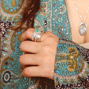 May include: A hand wearing a stack of silver rings with a turquoise gemstone. The rings are on a finger with a blue and green paisley patterned fabric in the background.