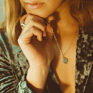 May include: A silver necklace with a teardrop-shaped pendant. The pendant has a light blue stone and is framed by a gold-colored metal. The necklace is worn against a teal velvet jacket.