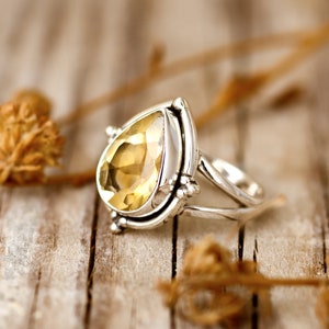 May include: A silver ring with a teardrop-shaped yellow gemstone set in a silver bezel. The ring has a delicate, intricate design with a split band.