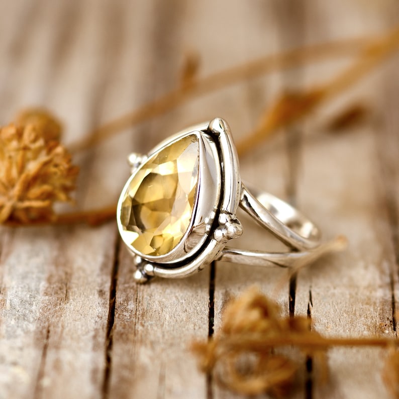 May include: A silver ring with a teardrop-shaped yellow gemstone set in a silver bezel. The ring has a delicate, intricate design with a split band.
