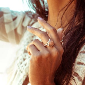 Op de afbeelding: Een zilveren ring met een zeshoekige, perzikkleurige edelsteen. De ring zit aan een vinger.