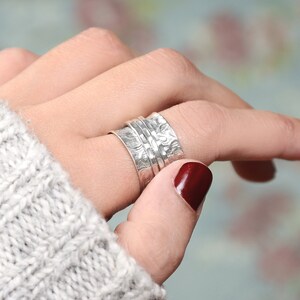 Puede incluir: Un anillo giratorio de plata con un dise&ntilde;o texturizado. El anillo se lleva en un dedo con esmalte de u&ntilde;as rojo.