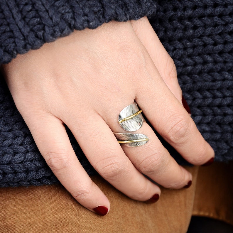 May include: A silver and gold ring with a leaf design. The ring is worn on the index finger of a hand.