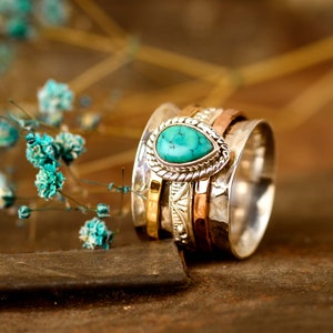 May include: A silver spinning ring with a turquoise gemstone set in a silver bezel. The ring is made of multiple bands of silver, gold, and copper.