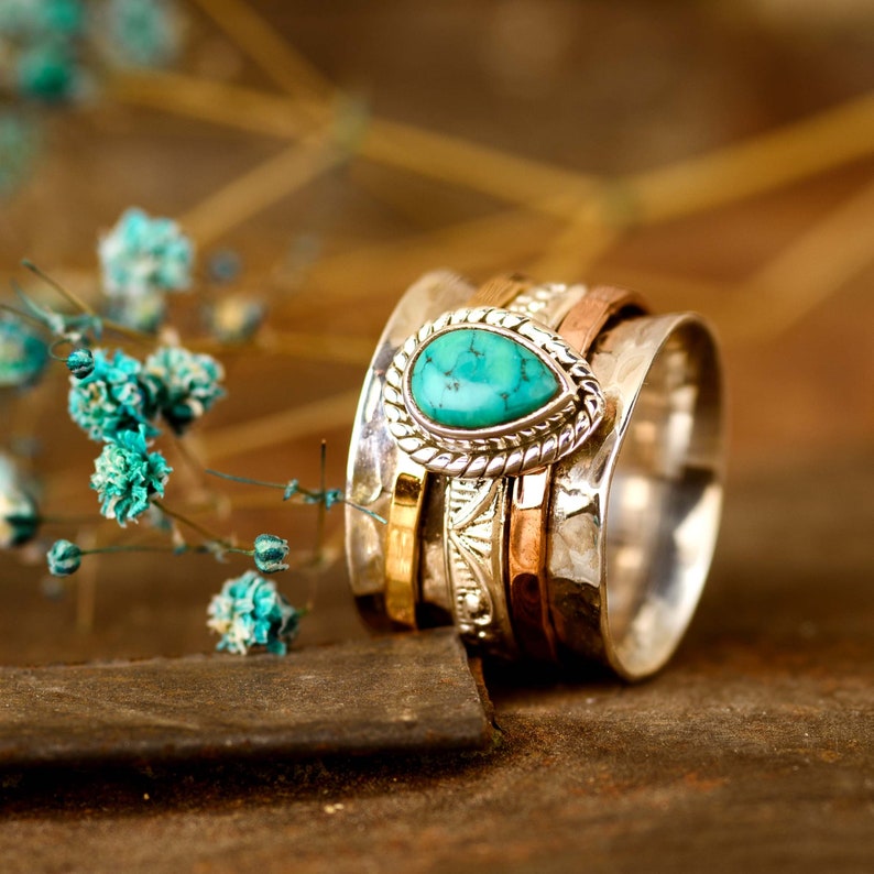 May include: A silver spinning ring with a turquoise gemstone set in a silver bezel. The ring is made of multiple bands of silver, gold, and copper.