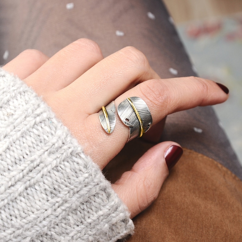May include: A silver ring with a gold accent, shaped like two overlapping leaves. The ring is worn on a finger.