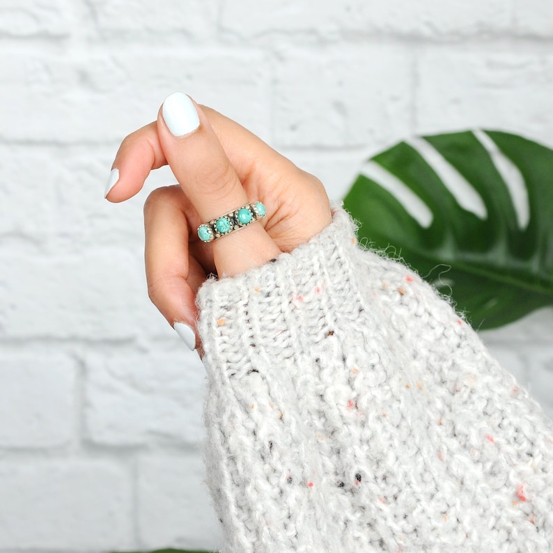 May include: A close-up of a hand wearing a silver ring with turquoise stones. The hand is wearing a white knitted sweater.