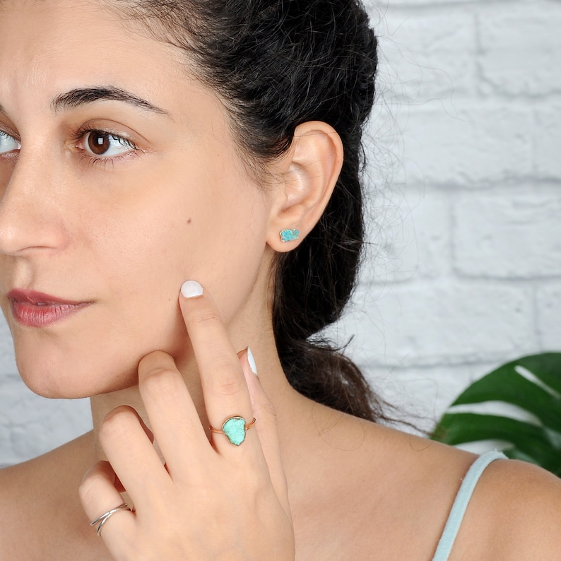 May include: A woman wearing a turquoise ring with a gold band. The ring features a unique, irregularly shaped turquoise stone. She is also wearing turquoise earrings.