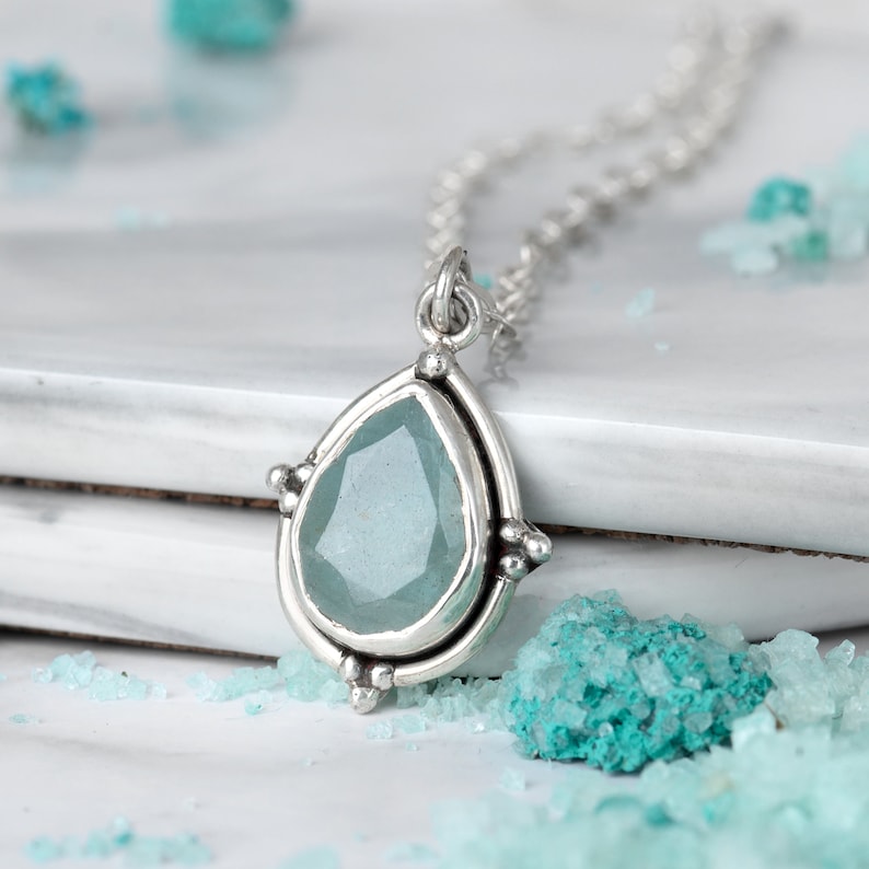 May include: A silver pendant necklace featuring a teardrop-shaped, pale blue gemstone. The pendant is set in a silver frame with decorative accents. The necklace is displayed on a white surface with scattered turquoise crystals.
