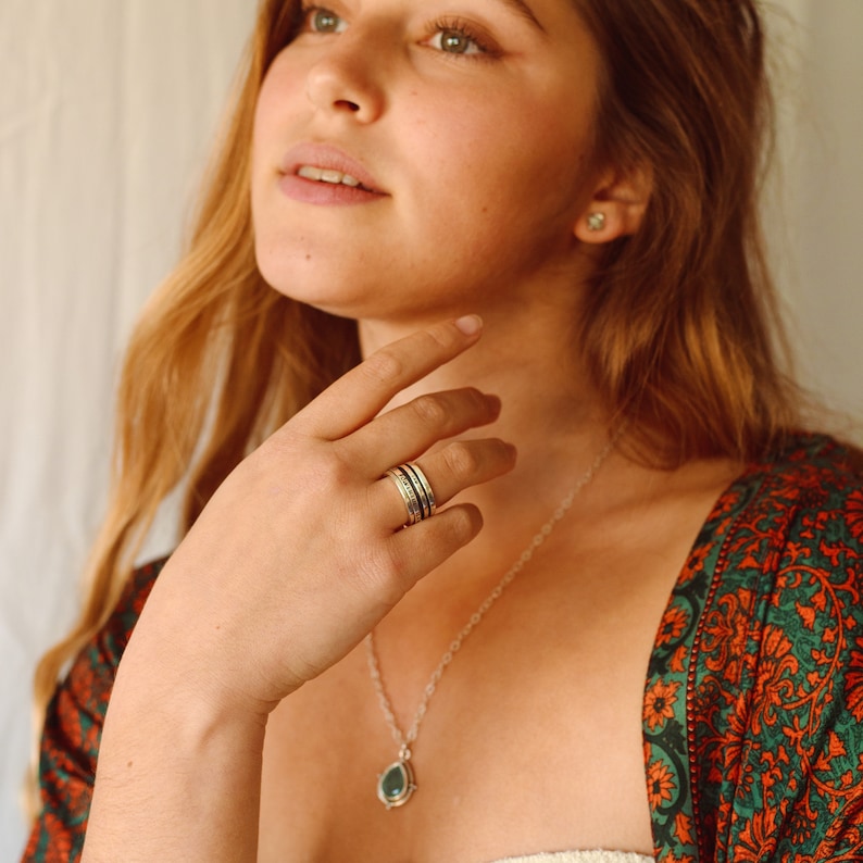 May include: A woman wearing a green and orange patterned shirt with a silver necklace and a silver ring on her finger. The necklace has a green gemstone pendant.