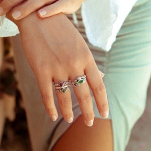 May include: Three silver rings with green and red gemstones. The rings are stacked on a finger.