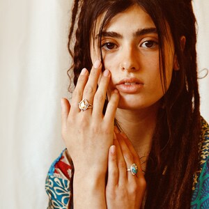 May include: A woman with long brown dreadlocks wears a colorful patterned shirt and two silver rings with gemstone accents. One ring features a teardrop-shaped yellow stone, and the other has a blue stone.