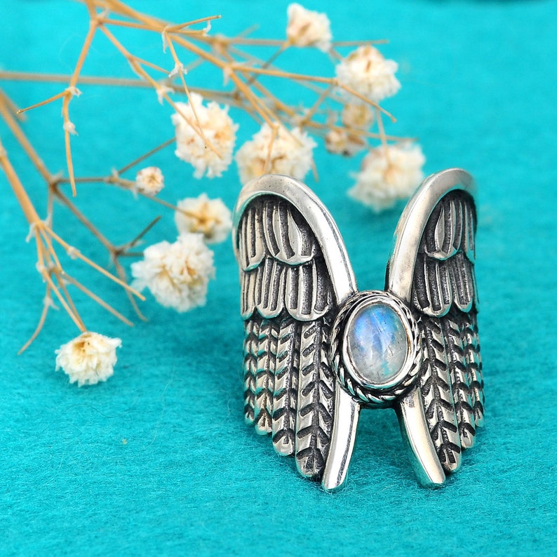 May include: A silver ring with a moonstone center and detailed wing designs on either side. The ring is on a turquoise background.