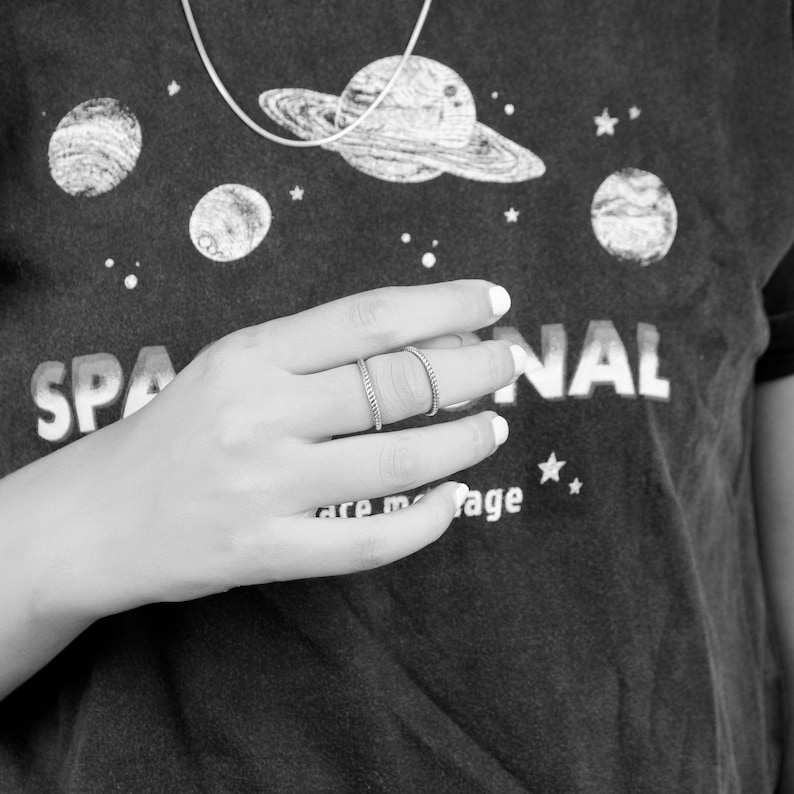 May include: A close-up of a hand wearing two silver rings. The hand is resting on a gray shirt with a space-themed design and the word "SPACENAL" printed on it.