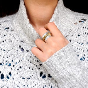 May include: A silver ring with gold wire accents is worn on a finger. The ring is on a hand, and the person is wearing a gray, open-knit sweater. A silver necklace is also visible.