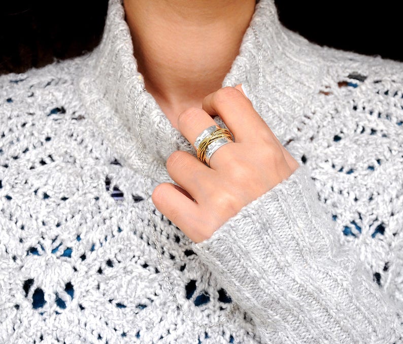 May include: A silver ring with gold wire accents is worn on a finger. The ring is on a hand, and the person is wearing a gray, open-knit sweater. A silver necklace is also visible.