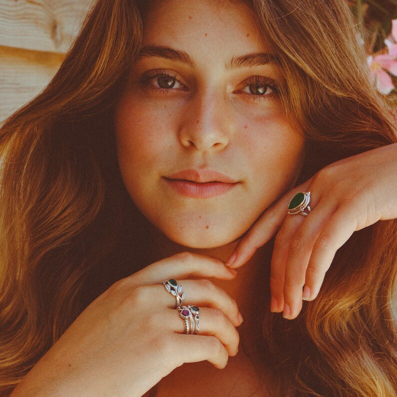 May include: A woman wearing several silver rings with green and red gemstones. The rings are on her fingers and are visible in the image.