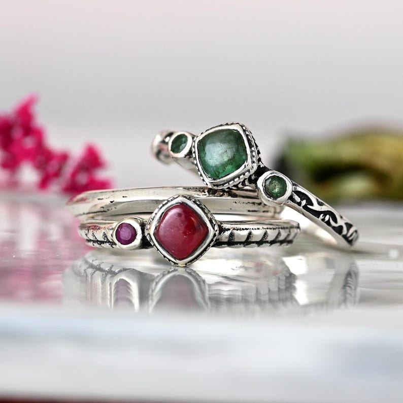 May include: Three silver rings with green and red gemstones. The top ring has a larger square green stone with two smaller green stones on either side. The middle ring has a square red stone with a smaller red stone on the side. The bottom ring is plain silver.