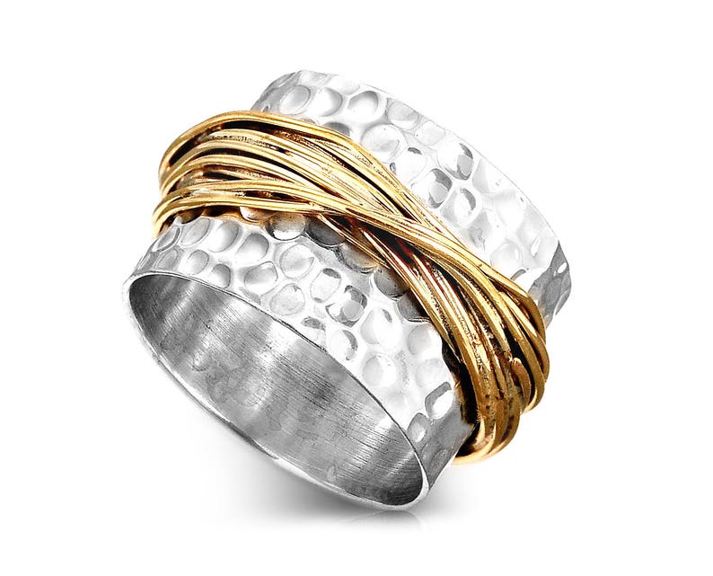 May include: A silver ring with a hammered texture and a gold-colored band wrapped around it. The ring has a wide band design and is set against a white background. The gold band adds a contrasting accent to the silver.