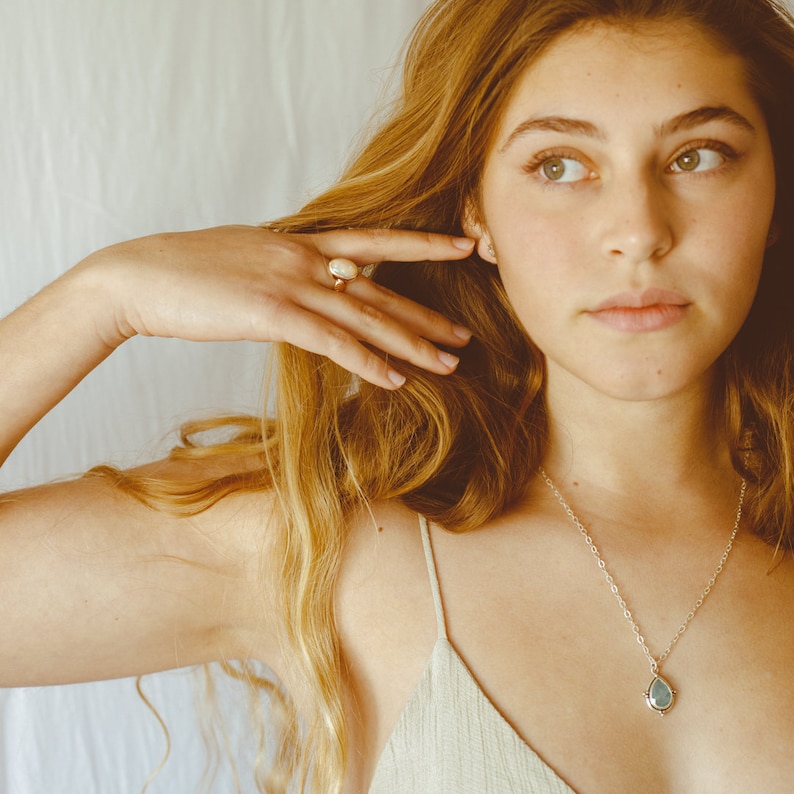 May include: A gold ring with a large, oval, white stone is worn on a finger. A silver necklace with a teardrop-shaped, light blue pendant hangs on a person. The person is wearing a light-colored top.
