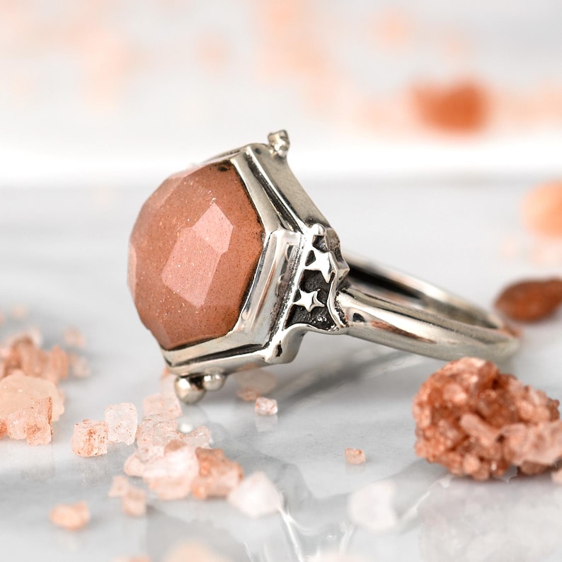 Op de afbeelding: Een zilveren ring met een grote, gefacetteerde, roze sunstone die in een zeshoekige bezel is gezet. De ring heeft een gedetailleerde band met stervormige accenten.