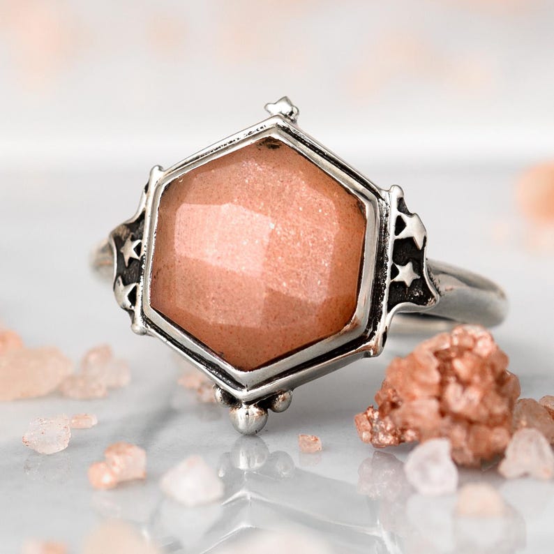 Op de afbeelding: Een zilveren ring met een zeshoekige roze sunstone edelsteen. De ring heeft een gedetailleerd ontwerp met sterren en kleine kralen.