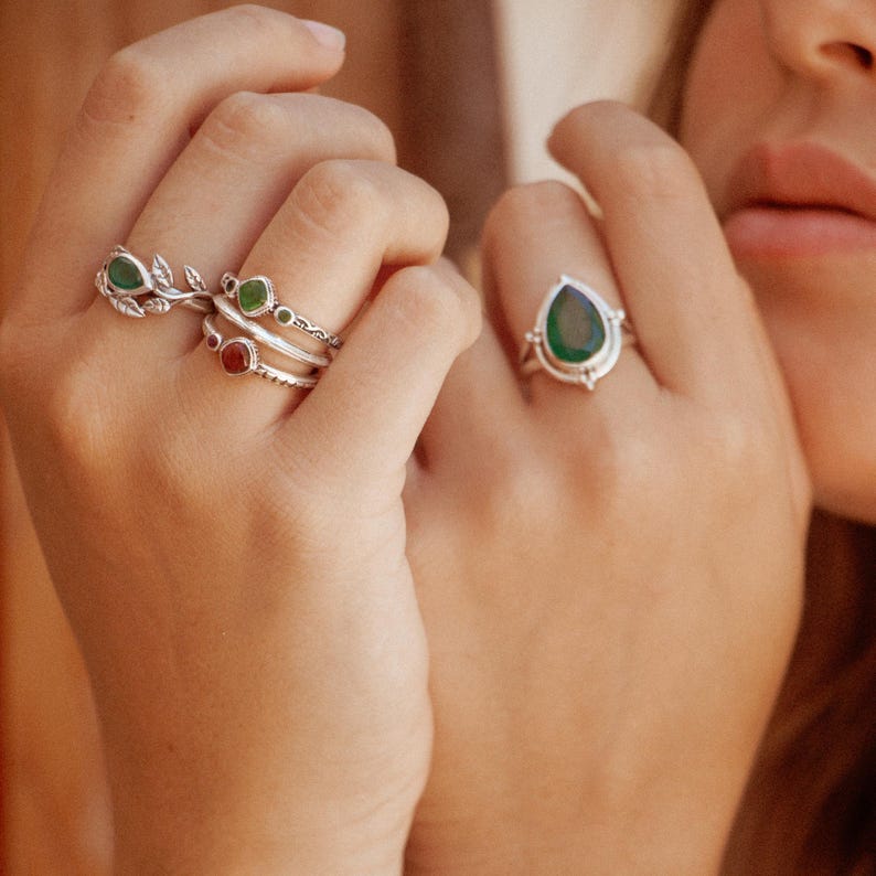 May include: A hand wearing a set of silver rings with green and red gemstones. The rings feature a leaf design, a small round stone, and a larger teardrop-shaped stone.