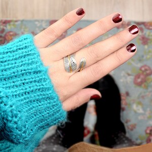 May include: A silver and gold ring with a leaf design. The ring is worn on the index finger of a hand with dark red nail polish.