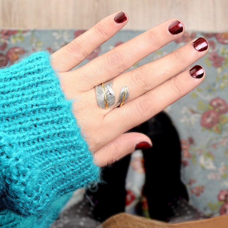 May include: A silver and gold ring with a leaf design. The ring is worn on the index finger of a hand with dark red nail polish.