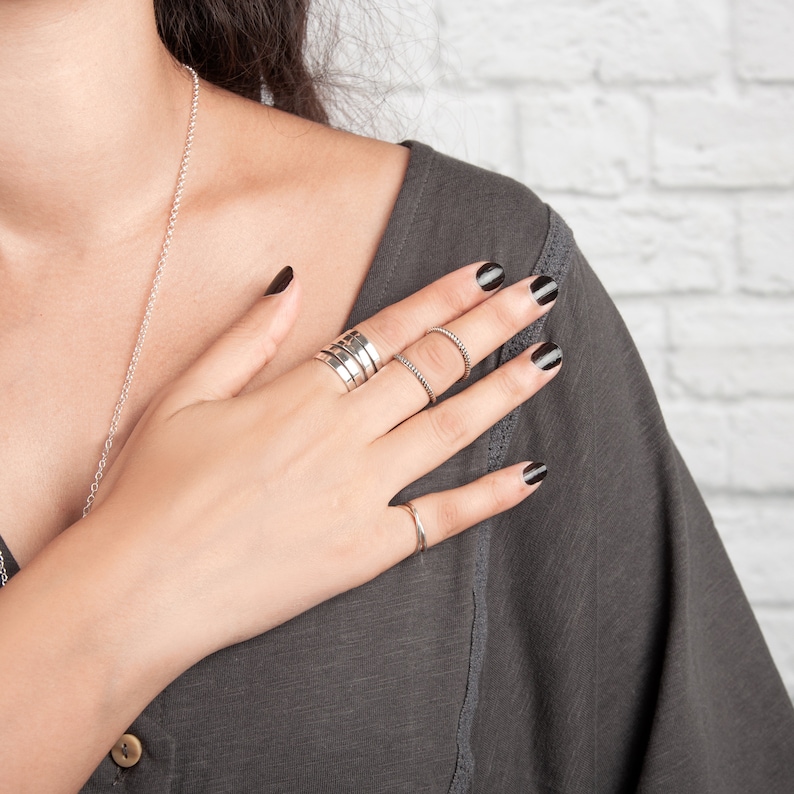 May include: A hand with black nail polish is wearing several silver rings. The hand is resting on a gray shirt.