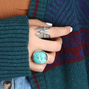 May include: Two silver rings with turquoise and moonstone gemstones. The ring on the index finger features a wing design. The ring on the middle finger has a round turquoise stone.