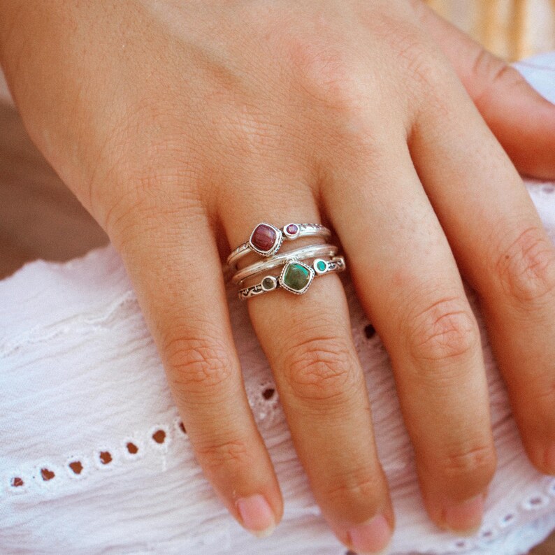 May include: Three silver rings with colorful gemstones. The top ring has a square red stone with two smaller red stones on either side. The middle ring has a square green stone with two smaller green stones on either side. The bottom ring has a silver design.