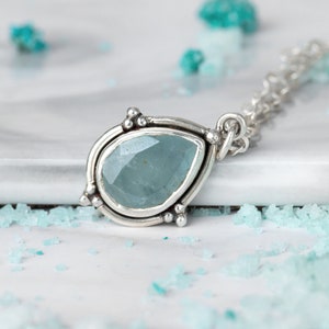 May include: A silver pendant necklace featuring a pear-shaped, light blue gemstone. The gemstone is set in a detailed silver bezel with small decorative silver beads. The necklace chain is also silver.