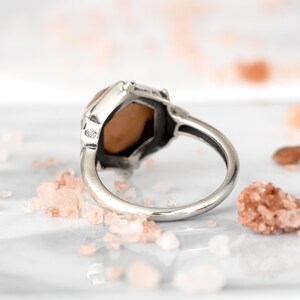 Op de afbeelding: Een zilveren ring met een zeshoekige edelsteen. De edelsteen is lichtroze.