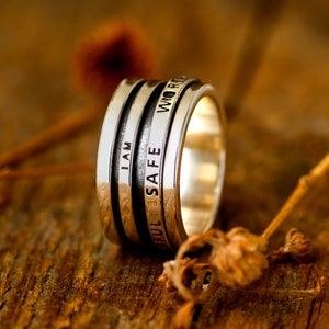 May include: A silver ring with three bands, each engraved with a different word: "I AM", "SAFE", and "WORTHY".