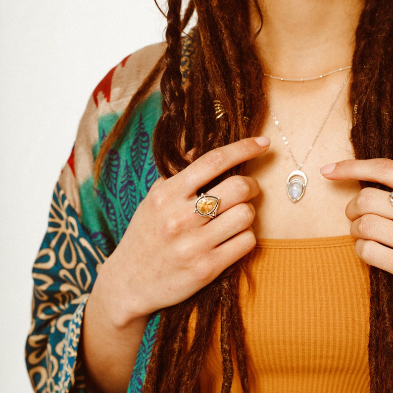 May include: A woman with long brown dreadlocks wears a colorful kimono and a silver necklace with a moonstone pendant. She is wearing a silver ring with a teardrop-shaped gemstone on her right hand.