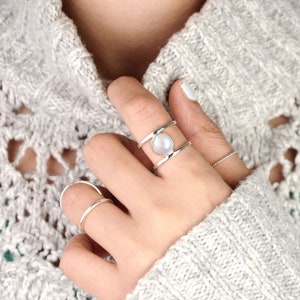 May include: A silver ring with a round, iridescent moonstone set between two parallel bands. The ring is worn on a finger, and a simple silver band is visible on another finger. The background is a light gray knitted sweater.