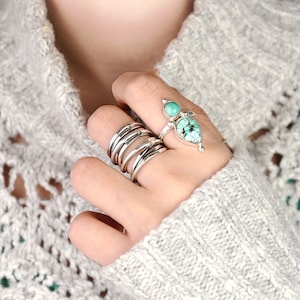 May include: A hand wearing a stack of silver rings and a turquoise gemstone ring with a teardrop-shaped stone.