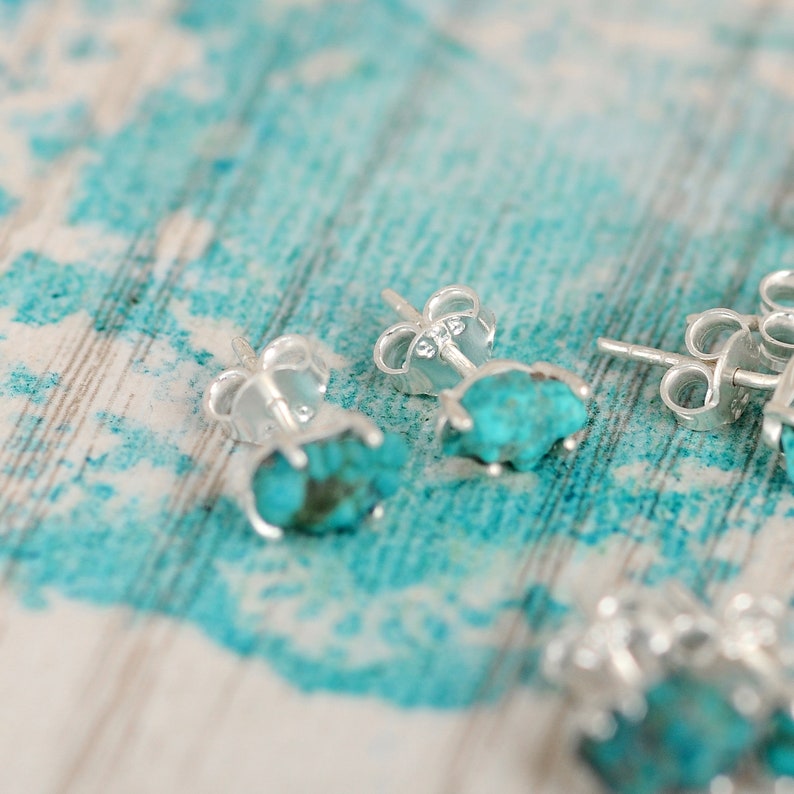 May include: Close-up of turquoise gemstone earrings with silver settings. The earrings are on a light blue and white wooden surface.