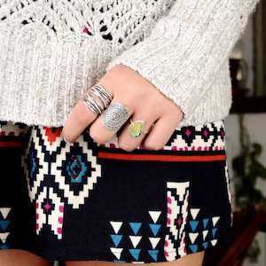 May include: A woman's hand wearing a silver ring with a decorative design and a green gemstone ring. The hand is wearing a grey sweater and a black and white patterned skirt.