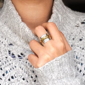 May include: A silver ring with a hammered texture and wrapped with gold-colored wire. The ring is worn on a finger, and the person is wearing a gray sweater. A silver necklace is also visible.