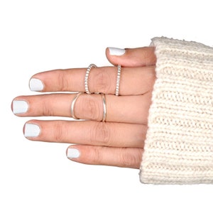 May include: A hand wearing four silver rings. Two rings are plain bands, and two are adorned with small, raised details. The hand is wearing a cream-colored sweater with white nail polish.