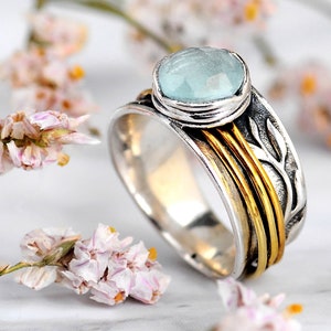 May include: A silver ring with a light blue gemstone. The ring features gold bands and a leaf design on the side. The gemstone is a light blue color and is set in a silver bezel. The ring is a piece of jewelry.