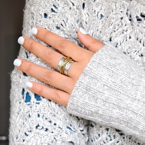 May include: A silver ring with a hammered texture and wrapped with gold-colored wire. The ring is on a hand with white nail polish, resting on a gray knit sweater. The ring is a piece of jewelry.