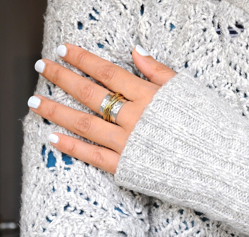 May include: A silver ring with a hammered texture and wrapped with gold-colored wire. The ring is on a hand with white nail polish, resting on a gray knit sweater. The ring is a piece of jewelry.