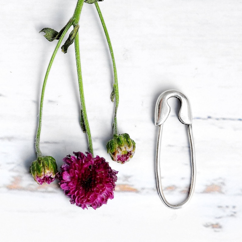 K&ouml;nnte beinhalten: Eine silberne Sicherheitsnadel mit abgerundeter Spitze und einer geschlossenen Schlaufe. Die Nadel liegt auf einer wei&szlig;en Oberfl&auml;che mit ein paar lila Blumen.