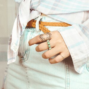 May include: A close-up of a person's hand wearing several silver rings with turquoise stones. The hand is wearing a light blue denim skirt and a white and light blue plaid shirt.