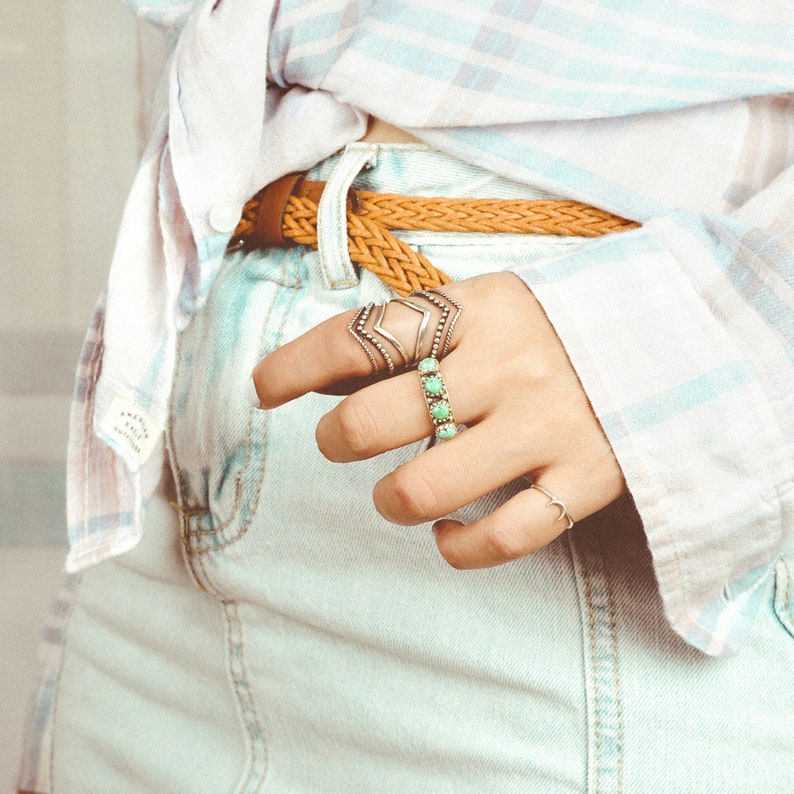 May include: A close-up of a person's hand wearing several silver rings with turquoise stones. The hand is wearing a light blue denim skirt and a white and light blue plaid shirt.