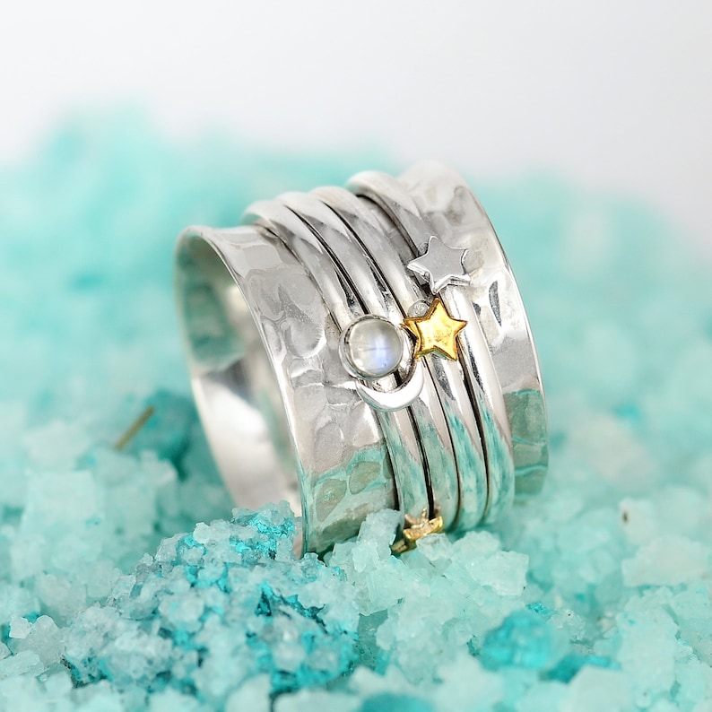 May include: A silver spinning ring with a moon and stars design. The ring is made of multiple bands and has a hammered texture. The moon is set with a clear gemstone.