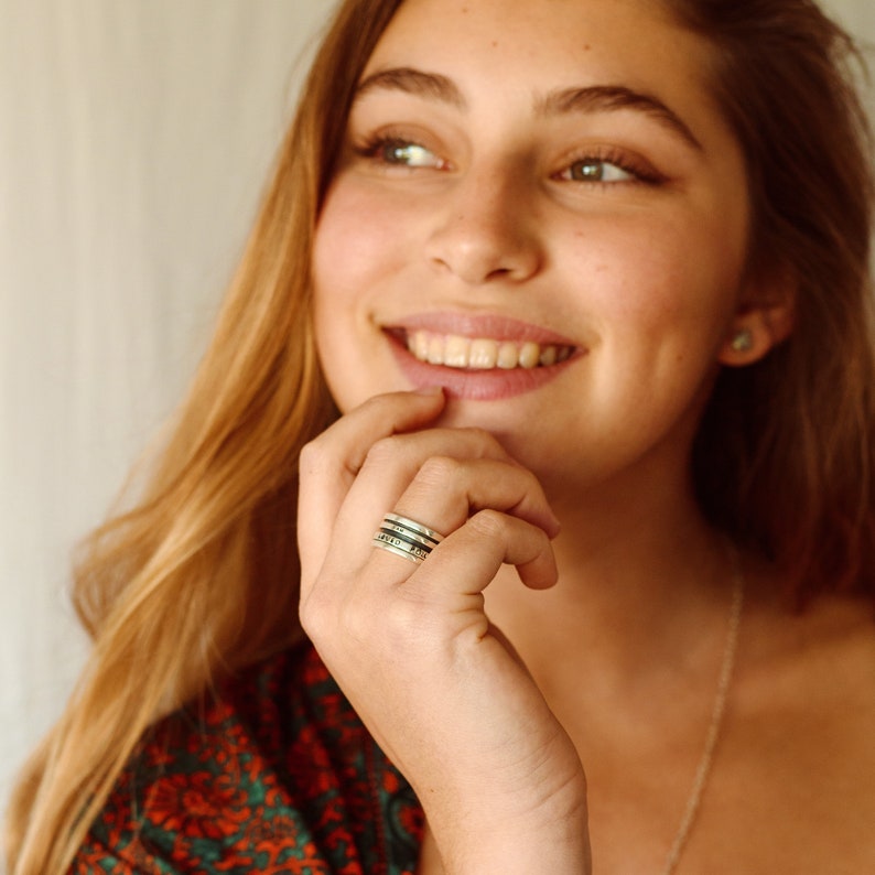 May include: A close-up of a woman's hand wearing a silver ring with multiple bands. The ring has an inscription on each band. The woman is smiling and looking off to the side.